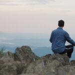 A man sitting on the rock and petting a dog surrounded by mountains under a cloudy sky